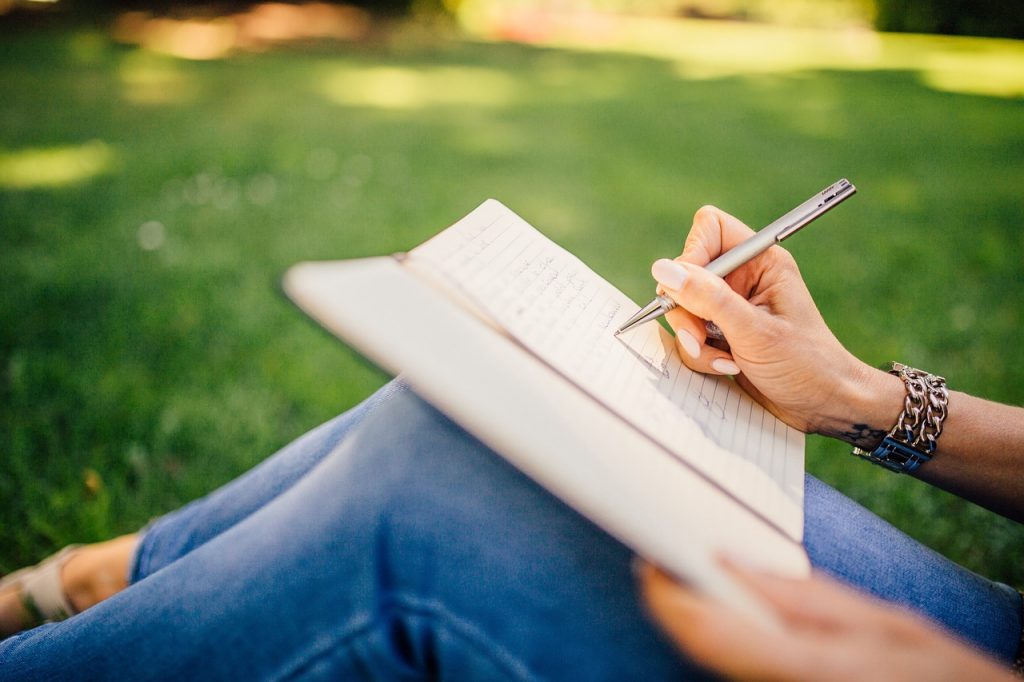 Person holding a pen, writing on a notebook on their lap. Sitting on grass, wearing blue jeans.